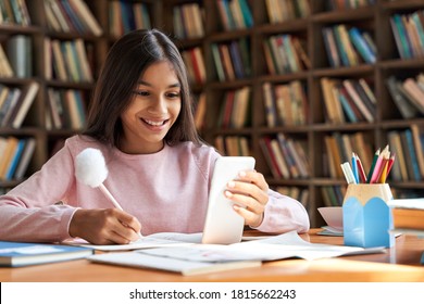 Indian Latin Child Preteen School Girl Holding Phone Distance Learning Class Using Mobile Application, Watching Online Lesson, Video Calling In App Making Notes Studying At Home, In Classroom.