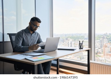 Indian latin focused financier sitting at desk doing paperwork working with bank taxes, loans, debit, credit operations using laptop near panoramic window in contemporary corporation office.
