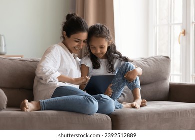 Indian little girl her mom enjoy digital tablet usage at home, sit on sofa look at screen choose goods, buy ecommerce services spend time on internet. Young generation use modern wireless tech concept - Powered by Shutterstock