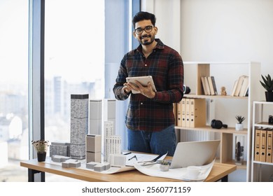 Indian male architect develops modern city project using digital tablet in creative workspace. Tall building models on desk symbolize innovative urban design. Age group young adult to middle-aged. - Powered by Shutterstock