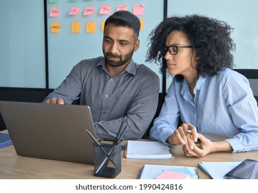 Indian male businessman executive manager mentor explaining working operations to female African American colleague coworker intern using laptop sitting in modern office conference room 