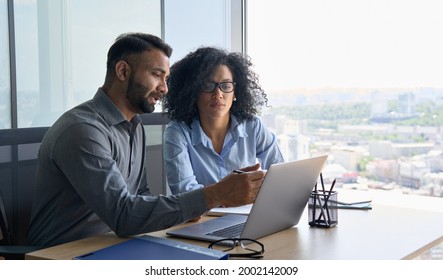 Indian male ceo executive manager mentor giving consultation financial operations to female African American colleague intern using laptop sitting in modern office near panoramic window 