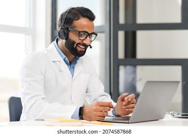 Indian Male Conference Speaker In A Medical Gown, Glasses And A Headset Sitting At The Desk And Teaching Or Talking Online With The Virtual Conference Participants, Healthcare Webinar Concept