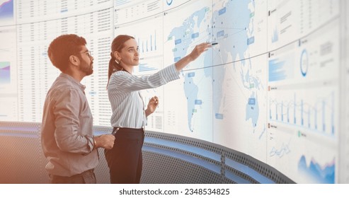 Indian Male Data Scientist And Caucasian Female Business Manager Talking In Front Of Big Digital Screen With Graphs, Charts And Map In Monitoring Office. Diverse Colleagues Discussing Objectives. - Powered by Shutterstock