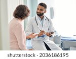 Indian male doctor consulting senior old patient filling form at consultation. Professional physician wearing white coat talking to mature woman signing medical paper at appointment visit in clinic.
