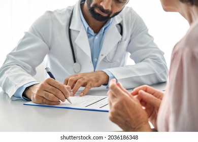 Indian Male Doctor Consulting Senior Old Patient Filling Medical Form. Professional Physician Writing Information In Clipboard Checking Examining Elderly Woman During Appointment Visit In Clinic.