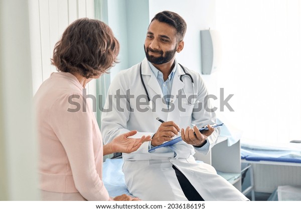 Indian male doctor consulting senior old patient
filling form at consultation. Professional physician wearing white
coat talking to mature woman signing medical paper at appointment
visit in clinic.