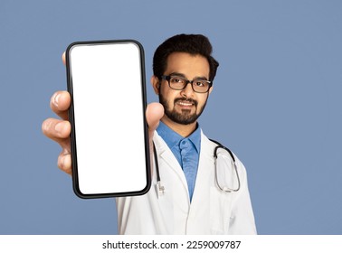 Indian Male Doctor Demonstrating Blank Smartphone Screen, Handsome Physician Man Showing Copy Space, Advertising Medical App While Standing Over Purple Studio Background, Creative Collage, Mockup - Powered by Shutterstock