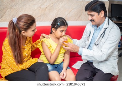 Indian Male Doctor Help Sick Child To Drink Juice While Mother Sitting Next To Him At Clinic Or Hospital. Children Healthcare Concept.