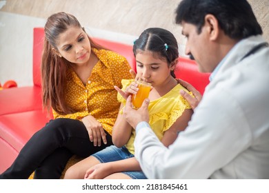 Indian Male Doctor Help Sick Child To Drink Juice While Mother Sitting Next To Him At Clinic Or Hospital. Child Healthcare Concept.