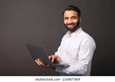 Indian Male Entrepreneur Carrying Laptop Isolated On Black, Bearded Businessman Using Laptop, Typing, Receiving Emails, Using New Computer App, Looking At The Camera And Smiles