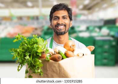 Indian Male Grocery Store Or Hypermarket Employee Holding Brown Paper Bag Of Vegetables With Friendly Smile Expression