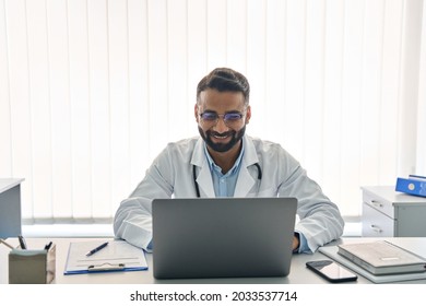 Indian male happy doctor medical worker in modern clinic wearing eyeglasses and white coat uniform using laptop computer having videocall chat, consulting remotely. Telemedicine health care concept.