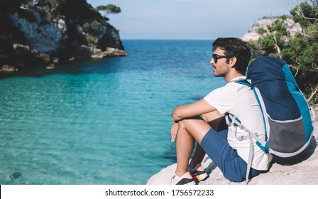 Indian male hiker resting at rocky abyss with breathtaking scenery views on turquoise water bay in Menorca, South Asian man with touristic backpack admire beautiful nature landscape during solo trip - Powered by Shutterstock