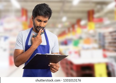 Indian male hypermarket or supermarket employee thinking as fingers touching chin with clipboard and market in hand