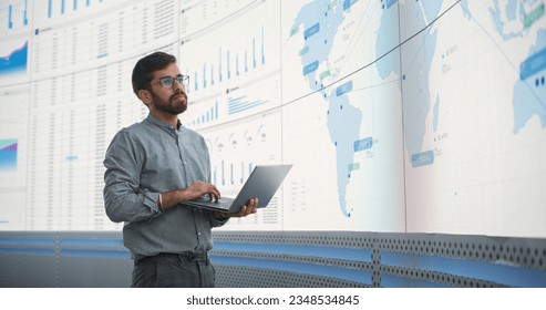 Indian Male Logistics Expert Holding Laptop Computer And Analyzing World Map On Big Digital Screen In Monitoring Office. Successful Man Developing New Efficient Routs For Global Product Distribution.