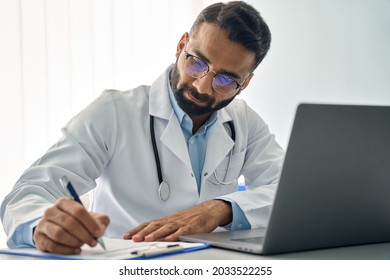 Indian male medical worker in modern clinic wearing eyeglasses and white doctor's coat using laptop computer writing health personal data, consulting remotely. Telemedicine healthcare concept. - Powered by Shutterstock