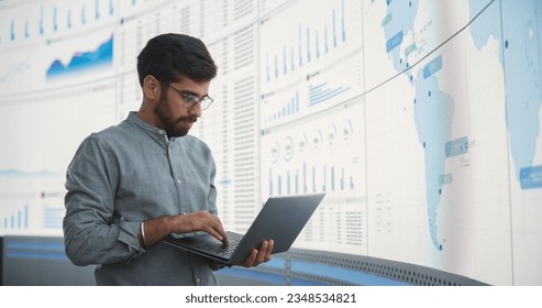 Indian Male Product Distribution Expert Holding Laptop Computer, Analyzing World Map On Big Digital Screen In Monitoring Office. Successful Man Looking For Efficient Routes For International Delivery.
