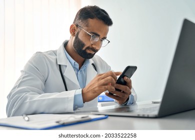 Indian male serious doctor medical worker in modern clinic wearing eyeglasses and white coat uniform using cell mobile smartphone apps, laptop computer. Medicine technologies health care concept. - Powered by Shutterstock