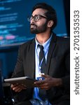 An Indian male software engineer or data scientist holds a tablet in a command center. He is asking a question about the AI code or cybersecurity data shown on the large screen.
