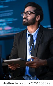 An Indian male software engineer or data scientist holds a tablet in a command center. He is asking a question about the AI code or cybersecurity data shown on the large screen.