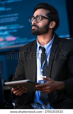 An Indian male software engineer or data scientist holds a tablet in a command center. He is asking a question about the AI code or cybersecurity data shown on the large screen.