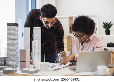 Indian man and African woman architects develop project for modern city. They collaborate at desk with building models and laptop.