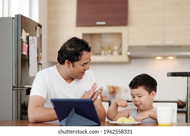 Indian Man Clapping To His Son Eating Plate Of Mashed Potato By Himself At Kitchen Table