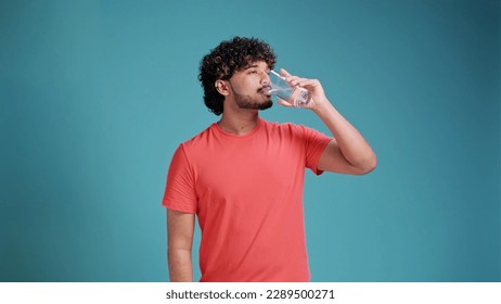 Indian man drinks water from a glass in coral t-shirt on blue studio background. - Powered by Shutterstock