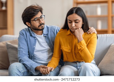 Indian man with glasses and a blue shirt sits on a couch beside a woman in a yellow top, comforting her as she appears upset. He holds her hand and gently rests his other hand on her shoulder - Powered by Shutterstock