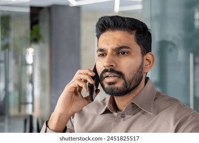 An Indian man looks tense and frustrated while talking on his mobile phone in a modern office setting, showing signs of anger and stress. - Powered by Shutterstock