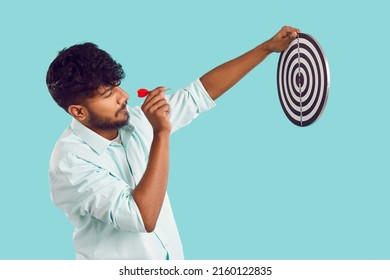 Indian man shooting at target. Profile view of young South Asian man in shirt standing isolated on blue background, holding dart board and aiming little red arrow at bullseye. Setting goal concept - Shutterstock ID 2160122835