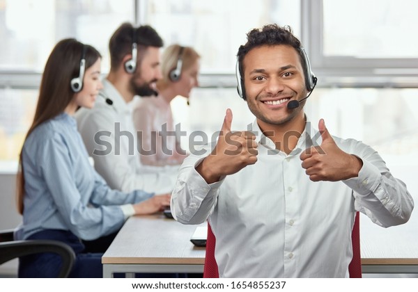 Indian man\
shows two thumbs up against bright office background. Happy call\
centre worker giving a thumbs up and looking to camera in front of\
working people in office\
backgorund.