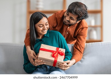 Indian man sitting on a couch is handing a gift to a woman next to him. The woman appears surprised and delighted as she receives the gift - Powered by Shutterstock