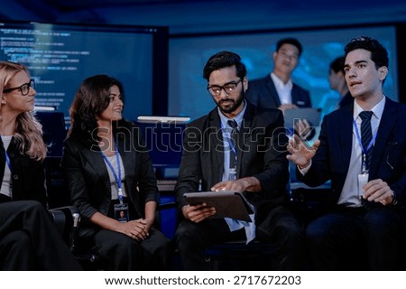 An Indian man with a tablet asks a question during a large team briefing. His diverse colleagues, including Indian and Caucasian women and men, listen in a high-tech conference room.