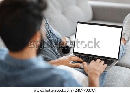 Indian Man Using Laptop Computer With Blank White Screen While Sitting On Couch At Home, Unrecognizable Male Working Online Or Browsing Internet Website, Over Shoulder View, Mockup With Copy Space