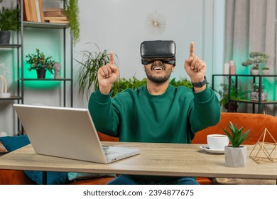 Indian man using virtual reality futuristic technology VR app headset helmet to play simulation 3D 360 online video game, watching film movie at modern home apartment. Guy in goggles sitting at table