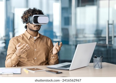 Indian man in VR glasses wearing sandy shirt while sitting by desktop with open laptop and raising hands in office. Creative digital designer using 3d innovation for working in cyber database. - Powered by Shutterstock