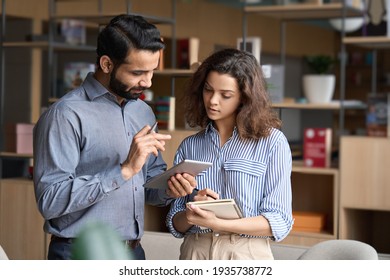 Indian manager holding digital tablet having discussion with latin employee meeting in office lobby. Two diverse professionals talking using pad discussing project strategy, checking corporate plan. - Shutterstock ID 1935738772