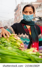 Indian Mature Woman Wearing Protective Face Mask And Buying Fresh Green Vegetables From Outdoor Market During The Third Wave Of Coronavirus, COVID-19 Pandemic Outbreak In Indian.    