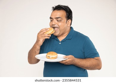 Indian Middle mature man holds and eating a hamburger deliciously. Concept of eating disorder and Relaxing with Eating junk food and unhealthy foods.  Burger is not helpful food with white background.