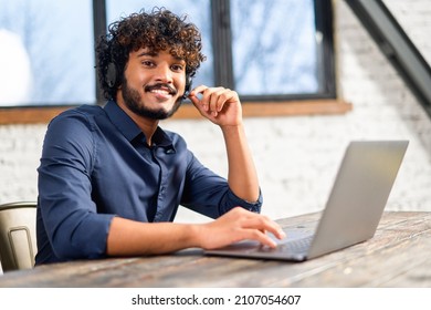 Indian Mixed Race Man Wearing Wireless Headset In Smart Casual Shirt Using Laptop While Sitting At The Desk. Inspired Hindu Male Employee In Headphones With Mic