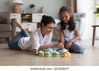 Indian mother and daughter play tea ceremony with cakes playthings seated and lying on warm floor in cozy living room, enjoy communication and playtime together at home. Toys store ad, hobby, leisure - Powered by Shutterstock