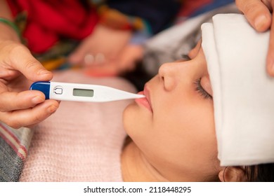Indian mother holding a thermometer in hand and measuring temperature of her ill daughter and sponging forehead to reduce the temperature down. She is having a high fever and lying in bed at home. 