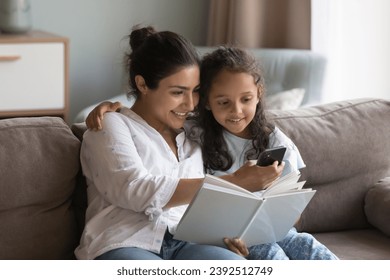 Indian mother and preschooler daughter sit on sofa at home holding storybook, distracted from reading use modern smartphone, search or buy books online via web store library, on-line bookshop concept - Powered by Shutterstock