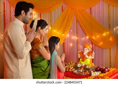 Indian nuclear family in traditional dress celebrating Ganesh chaturthi. The family is worshiping Lord Ganesh on Ganesh Chaturthi by folding their hands and eyes closed - Prayer