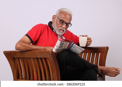 Indian Old Man Reading Magazine While Sitting On Chair With A Coffee Mug.