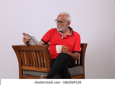 Indian Old Man Reading Magazine While Sitting On Chair With A Coffee Mug.