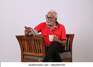 Indian Old Man Reading Magazine While Sitting On Chair With A Coffee Mug.