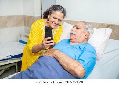 Indian Old Senior Patient In Hospital Bed Using Smart Phone Doing Video Call To Descendant Relatives With Wife Standing By Side - Elderly Medical And Healthcare Concept